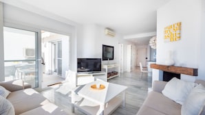 Un salon moderne avec un téléviseur à écran plat, une table basse blanche et un canapé. On y trouve une cheminée avec un manteau de cheminée et une œuvre d’art accrochée au mur.