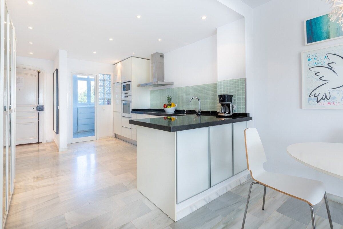 Una cocina moderna con una isla central, un comedor con una mesa y silla blancos, y vista del espacio de estar contiguo.