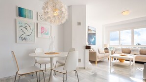 Un salon moderne avec une table à manger ronde, des chaises blanches, un canapé beige et une table basse. Les murs sont ornés d’œuvres d’art encadrées et une grande fenêtre laisse entrer la lumière naturelle.