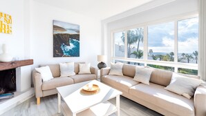 Un salon moderne avec un canapé beige, une table basse blanche et une cheminée. Une grande fenêtre offre une vue sur les palmiers et l’océan.