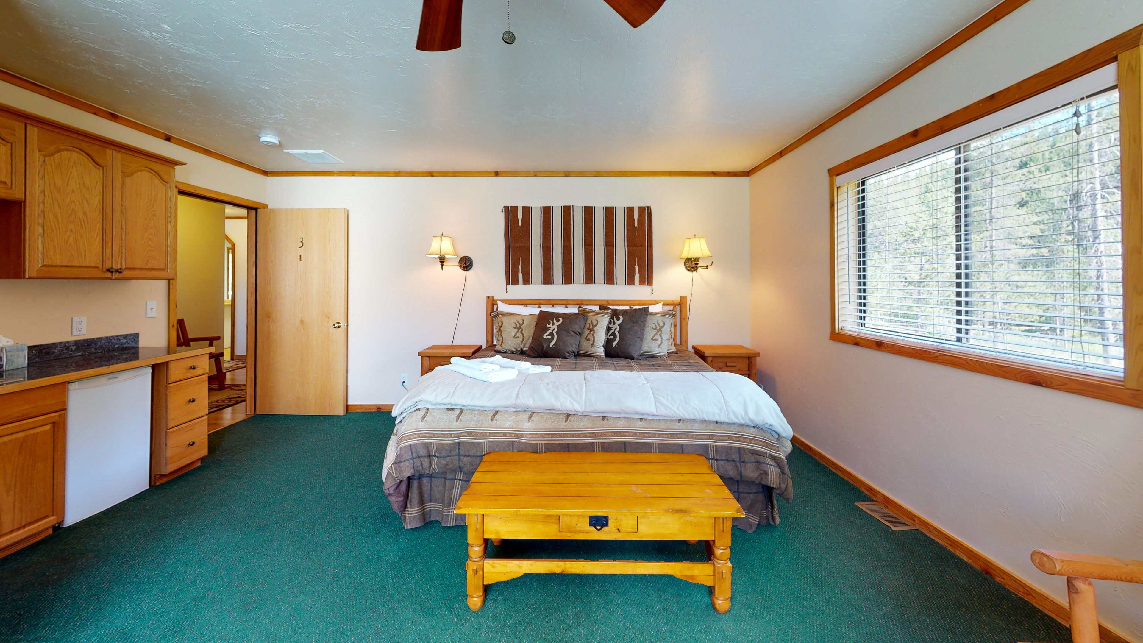A bedroom with a bed, bedside lamps, a wooden nightstand, a window with blinds, and a green carpet.