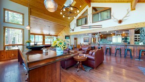 A spacious living area with a wooden floor, a central island with a sink, a sofa, and a dining table. There are multiple windows providing natural light and a view of the outdoors. - 40-acre haven on the banks of the Fall River (Ashton)