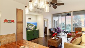 Ein geräumiges Wohnzimmer mit einem Holzesstisch, einem grünen Schrank und Korbmöbeln. Ein großer Fernseher ist an der Wand befestigt, und große Fenster bieten einen Blick ins Freie.