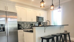 Una cocina moderna con armarios blancos, un refrigerador de acero inoxidable, un microondas y una cafetera. Hay dos taburetes negros y un ventanal que proporciona luz natural.