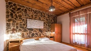 A bedroom with a stone wall, wooden ceiling, a bed with white bedding, a wooden wardrobe, and a window with curtains.