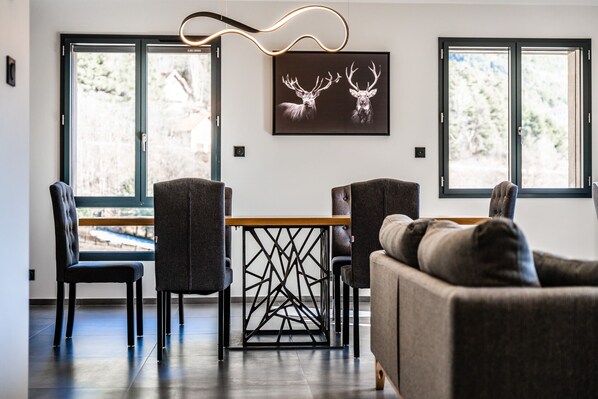 A modern dining area with a table and chairs, large windows, and a wall-mounted light fixture. - Charming mountain apartment (Saint-Chaffrey)