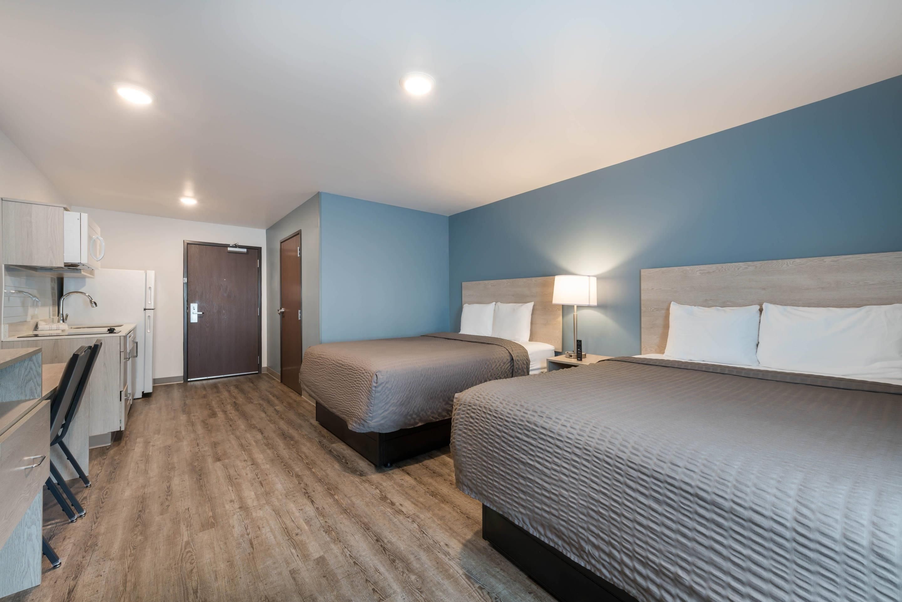 A hotel room with a kitchenette, two beds, a desk, and a bathroom.