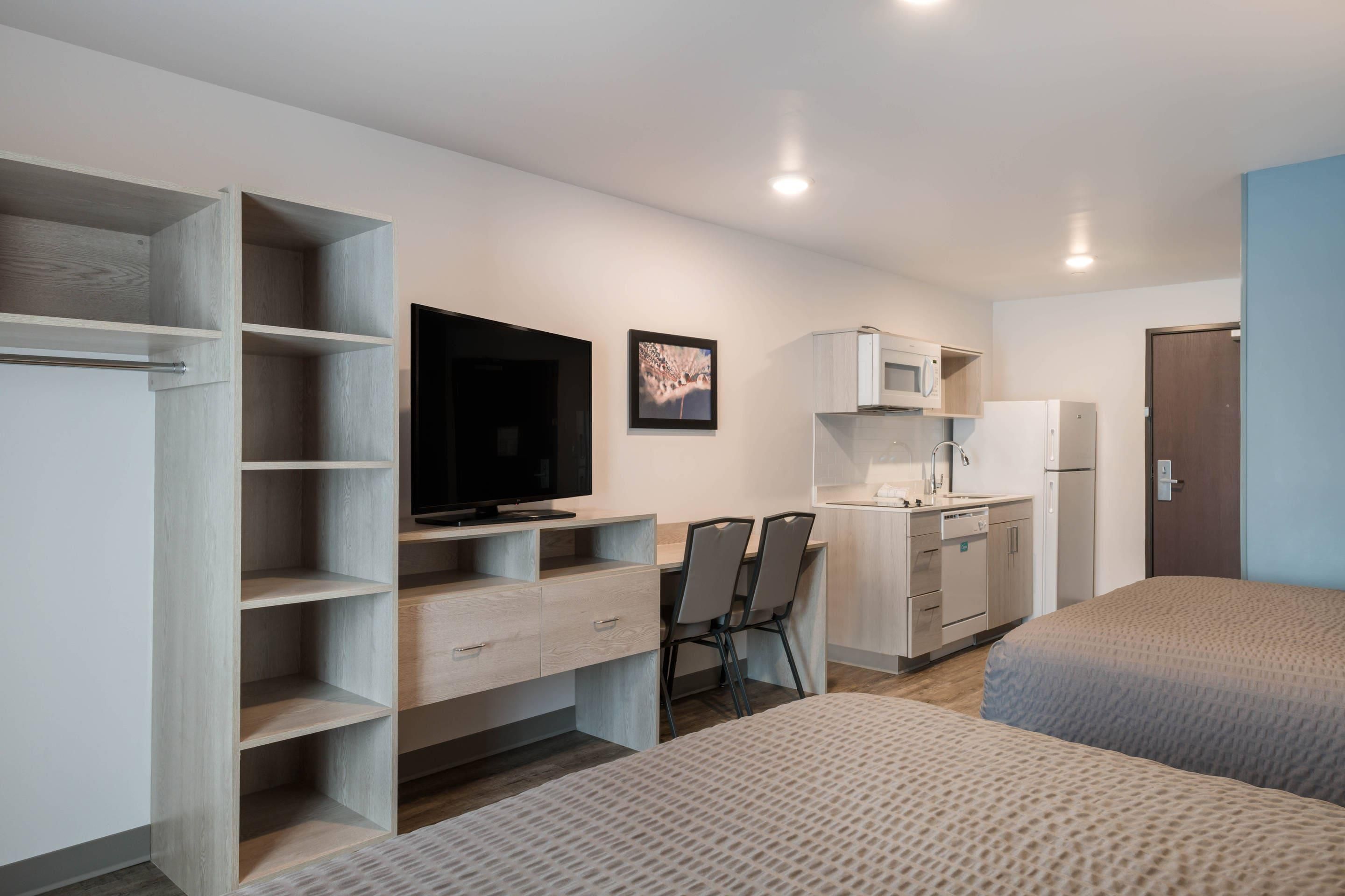 A compact hotel room with a kitchenette, a bed, a desk with a chair, and a television.