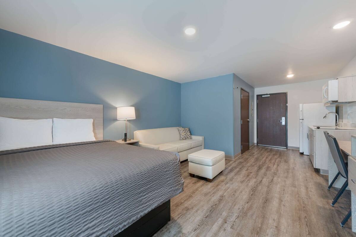 A modern bedroom with a large bed, a white sofa, a small table, and a kitchenette area.