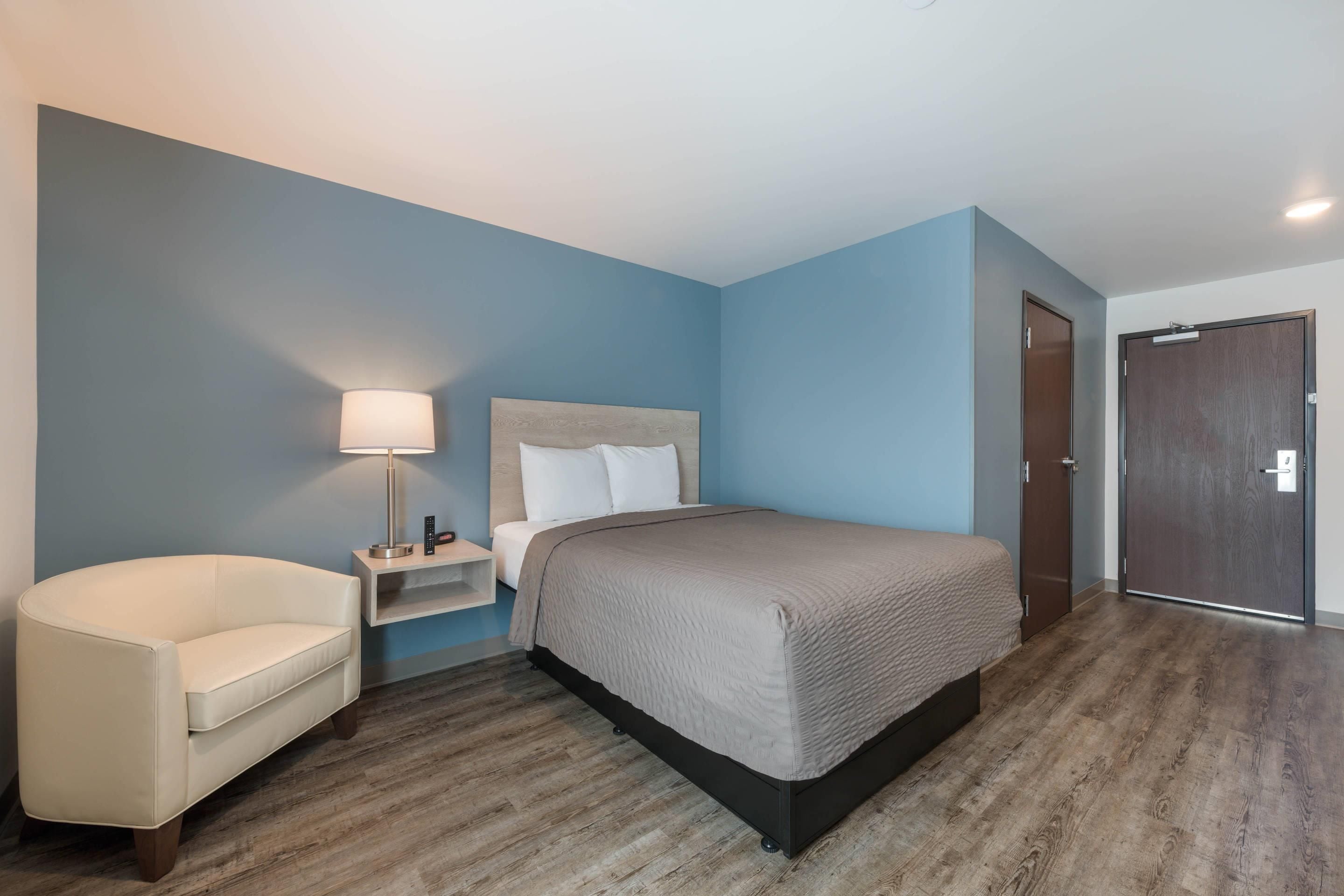 A hotel room with a bed, a chair, a nightstand, and a door.