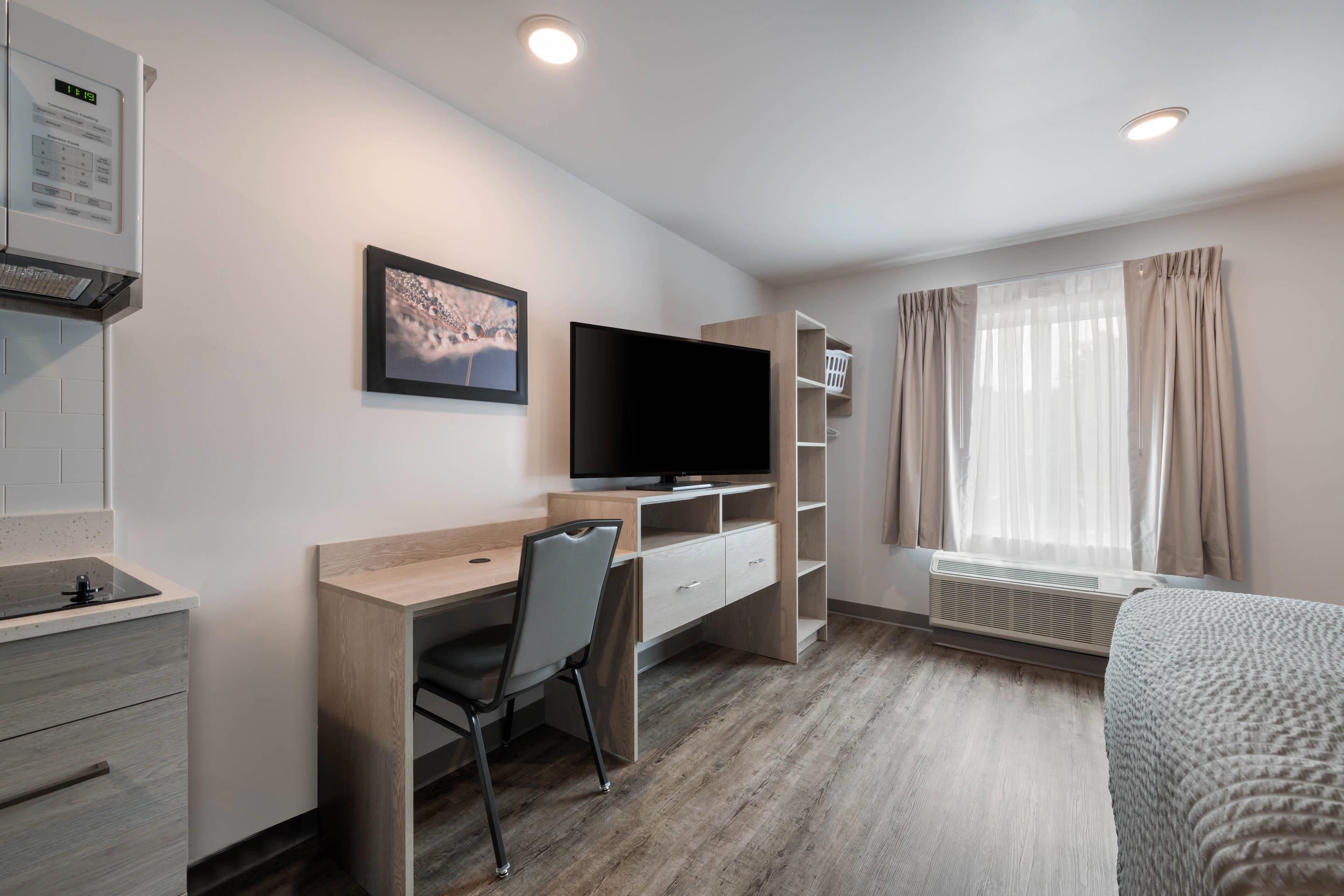 A modern hotel room with a wooden desk, a flat-screen TV, a bed, and a window with curtains.