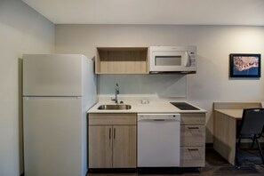 A compact kitchen with a refrigerator, microwave, sink, dishwasher, and a small dining area with a chair. - WoodSpring Suites Concord-Charlotte Speedway (Concord)