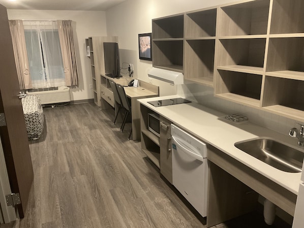 A modern kitchen with a dishwasher, sink, and a built-in oven. There is a window with curtains and a small table with a patterned tablecloth. - WoodSpring Suites Concord-Charlotte Speedway (Concord)
