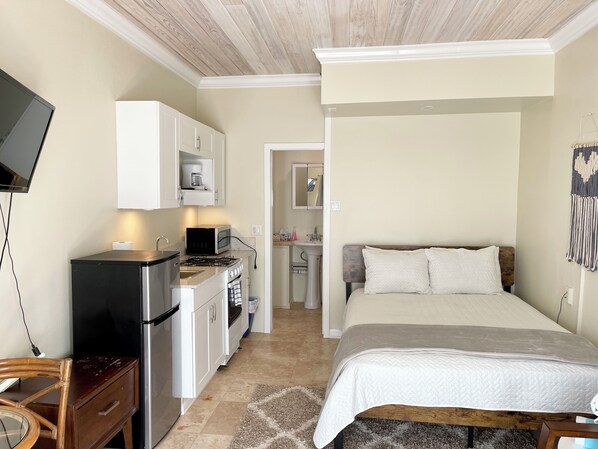 Un dormitorio compacto con cama, mesita de noche y un escritorio pequeño con silla. Una kitchenette con estufa, microondas y armarios se encuentra adyacente a la habitación.