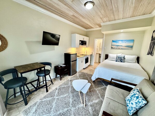 Une chambre d’hôtel compacte dotée d’une kitchenette, d’un lit donnant sur la plage, d’un petit coin repas et d’un téléviseur fixé au mur.