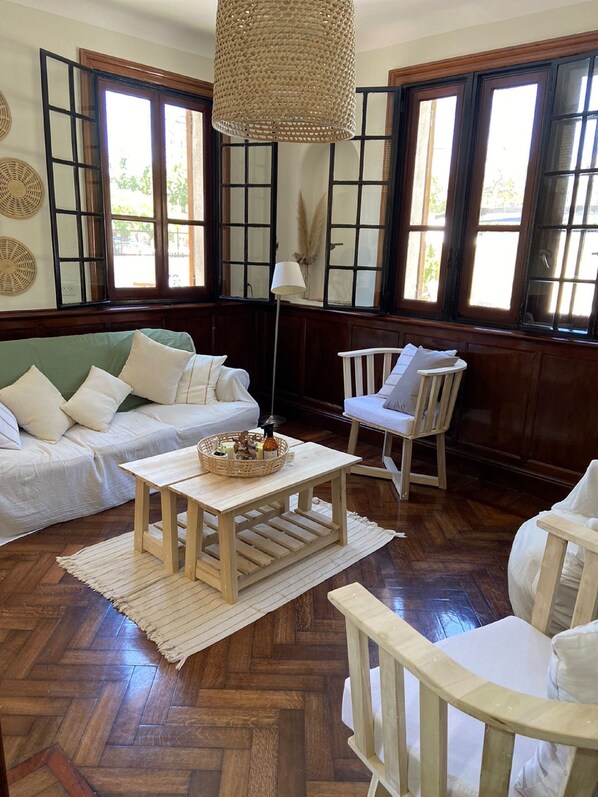 Quarto com piso de madeira em padrão herringbone, um sofá branco, uma cadeira branca, uma mesa de madeira pequena e um pendente tecido.