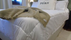 Una cama con ropa de cama blanca, una manta tejida y un almohadón con la palabra 'relax'.