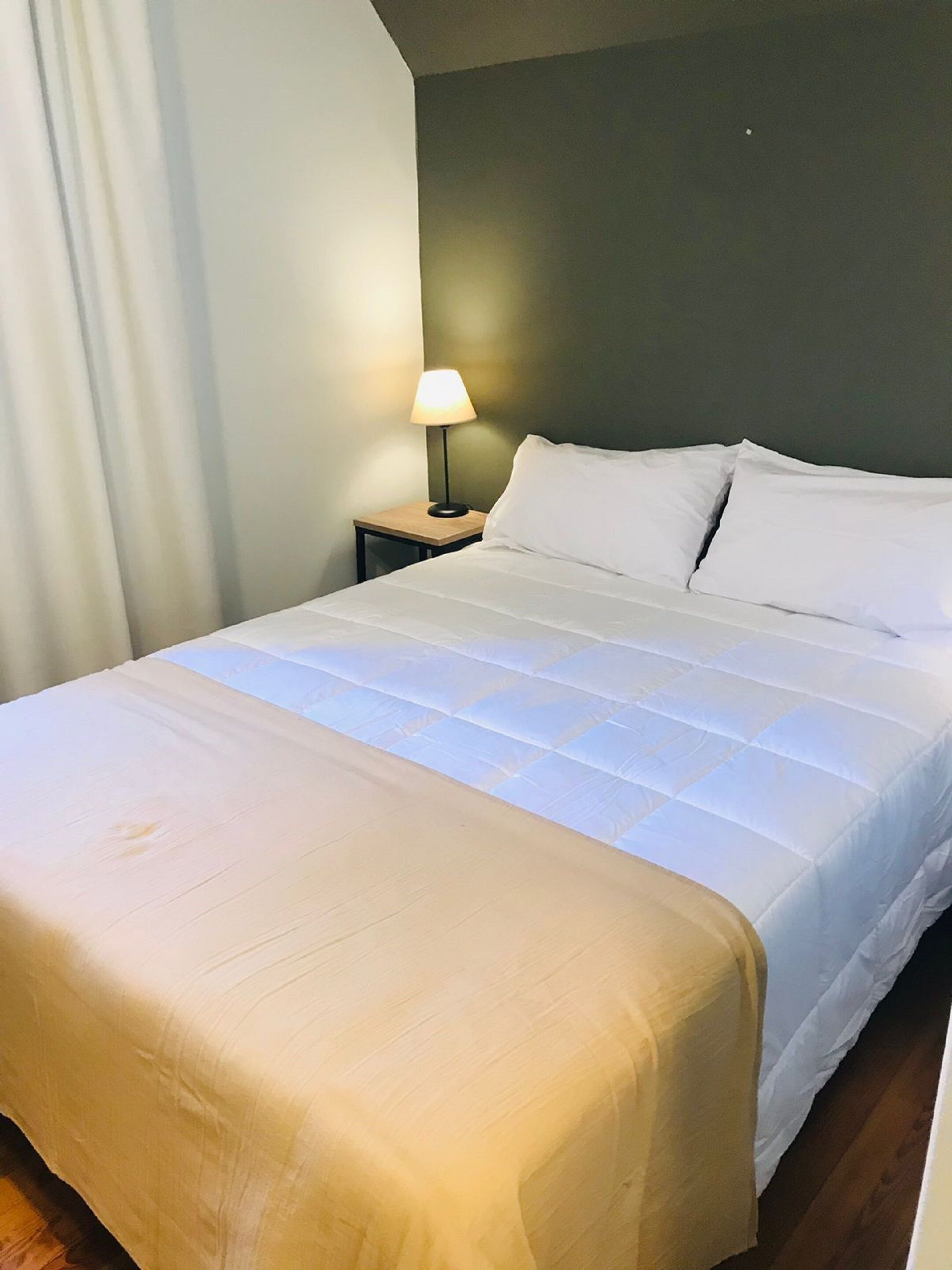 A neatly made bed with a white duvet and pillows, a bedside lamp, and a wooden nightstand.