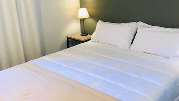 Uma cama bem arrumada com edredom branco e almofadas, uma luminária de cabeceira e uma mesinha de madeira.