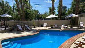 Una piscina con sillas de descanso y sombrillas, rodeada de árboles y una valla.