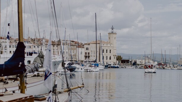 Un port de plaisance avec des bateaux amarrés, un bâtiment imposant surmonté d’une tour, et une eau calme dans laquelle se reflètent les bateaux.
