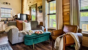 A cozy living room with a sofa, armchair, and ottoman, wooden walls, and a window with blinds.