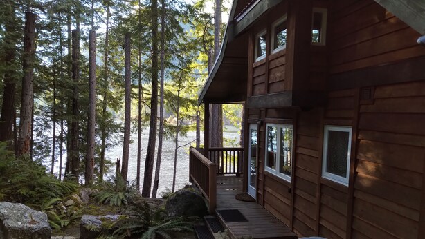A wooden cabin with a porch overlooking a forested area and a body of water. - The Cabin at Moon Dance (Madeira Park)