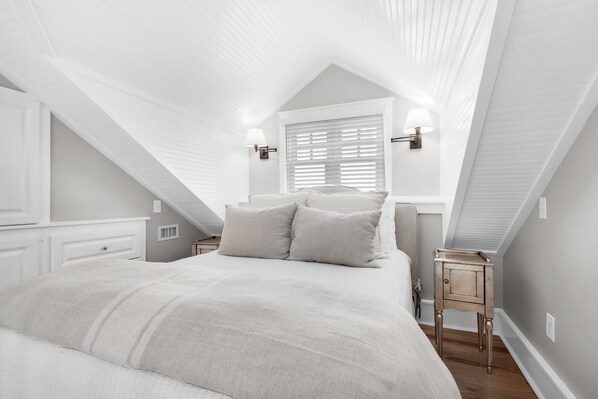 Ein Schlafzimmer mit schrägem Dach, einem Bett mit Kissen, einem Nachttisch, einer Lampe und einem Fenster mit Jalousien.
