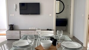 Un coin repas moderne avec une table en verre dressée pour six personnes, des assiettes blanches et des verres à vin transparents. Un téléviseur à écran plat est fixé au mur au-dessus de la table.