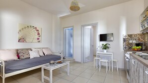 A modern living room with a sofa, coffee table, and a TV mounted on the wall. There is a kitchen area with a sink and a fruit bowl. - Cozy Vintage Style Apartment Shared Pool RU (Malevizi)