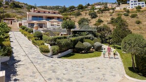 Un camino pavimentado que conduce a una propiedad en la ladera de una colina, con una casa moderna, rodeada de jardines cuidados y árboles.