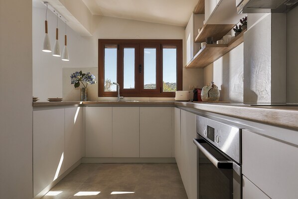 A modern kitchen with white cabinetry, a stainless steel oven, a window with a view, pendant lights, and a vase with flowers.