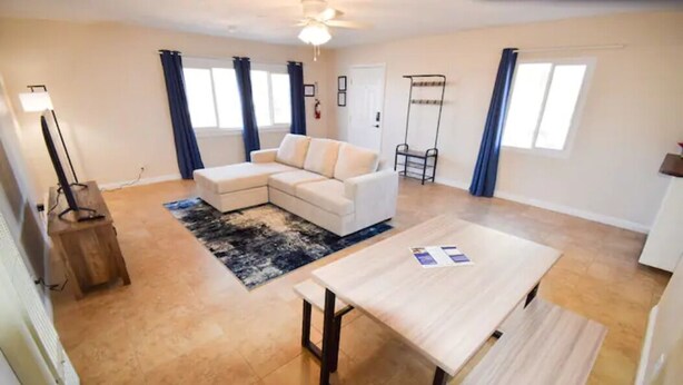 A living room with a white sofa, a coffee table, and a rug. There are two windows with curtains, a ceiling fan, and a small wooden table.