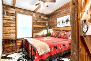 A bedroom with a wooden wall, a bed with a red blanket, a ceiling fan, and a window.