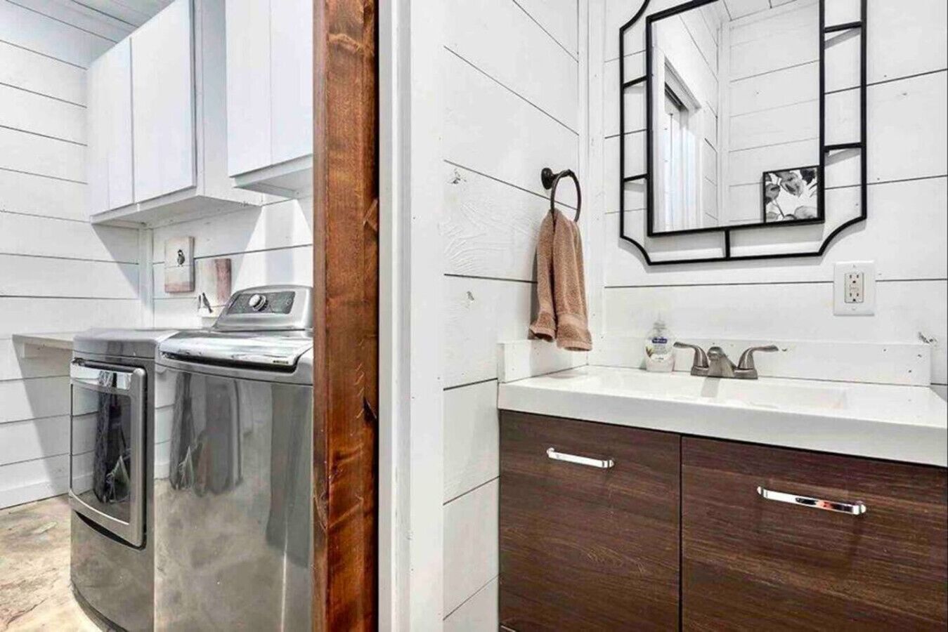 Une salle de bain moderne avec un lave-linge et un sèche-linge en acier inoxydable, un lavabo blanc, des meubles en bois foncé et un miroir avec un cadre noir.