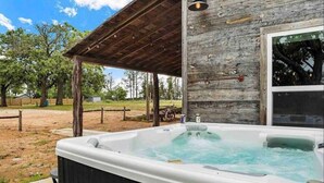 A hot tub outdoors with a wooden deck and a rustic building in the background. - The Sanctuary at Fredericksburg (Gillespie County)