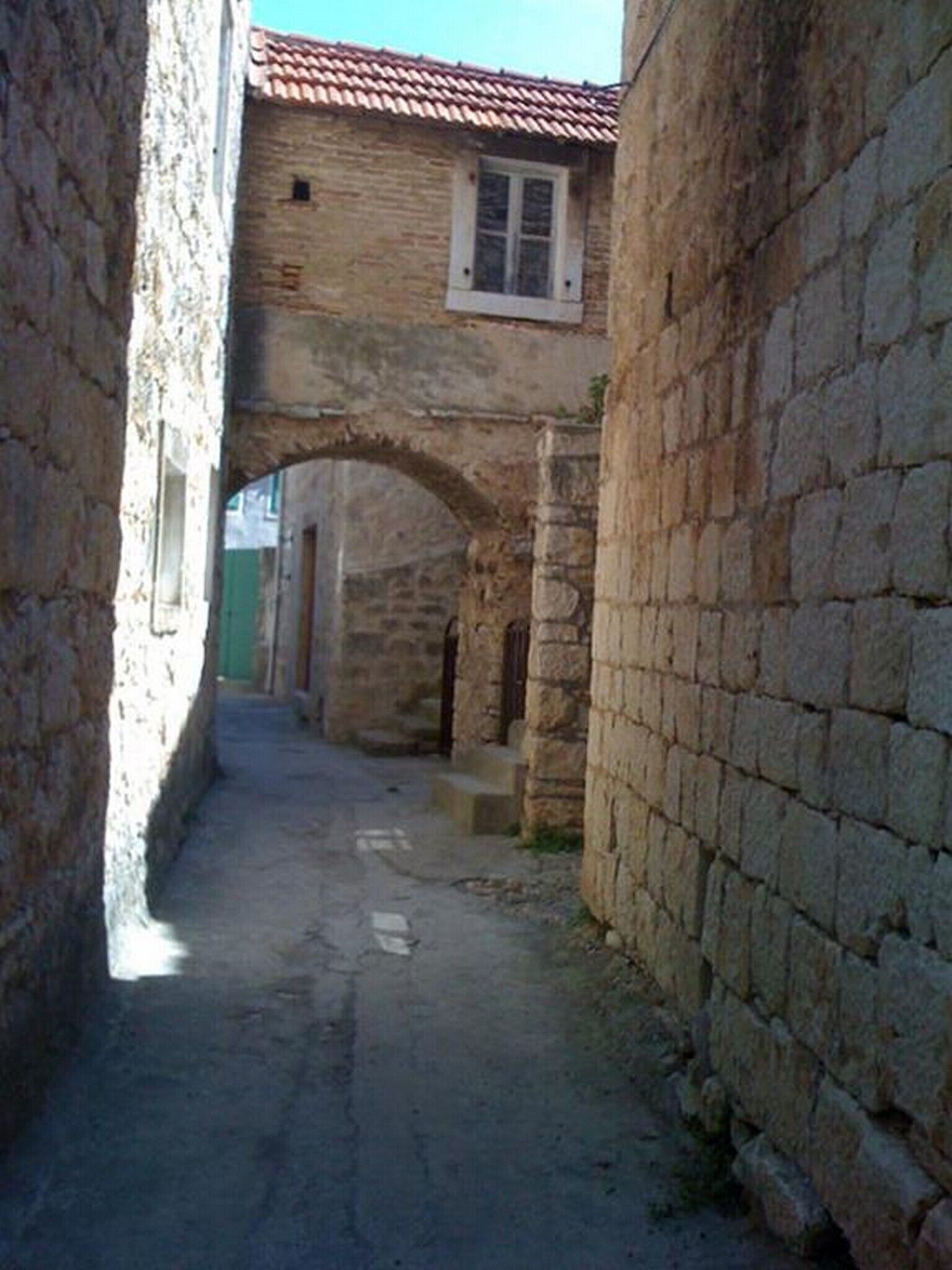 A narrow stone alleyway with a rustic building featuring a tiled roof and a window.
