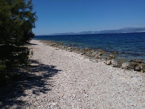 Ein Kiesstrand mit klarem, blauem Wasser und Bergen im Hintergrund.