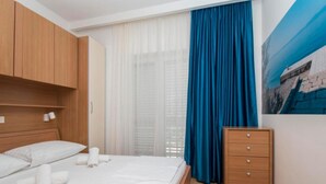 A neatly made bed with white linens and pillows, a wooden dresser with drawers, a large window with sheer white curtains, and a blue curtain. - Apartments Antonio - One Bedroom Apartment with Balcony - 1 (Tucepi)