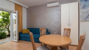 A room with a round wooden table and four chairs, a blue sofa with yellow cushions, a sliding glass door, and a grey textured wall. - Apartments Antonio - One Bedroom Apartment with Balcony - 1 (Tucepi)