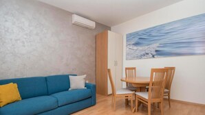 A living room with a blue sofa, wooden dining table, and chairs. There is a painting on the wall and an air conditioning unit mounted on the wall. - Apartments Antonio - One Bedroom Apartment with Balcony - 1 (Tucepi)