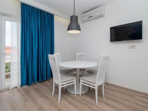 A room with a round white table, four white chairs, a black pendant light, a flat-screen TV on the wall, and an air conditioning unit.