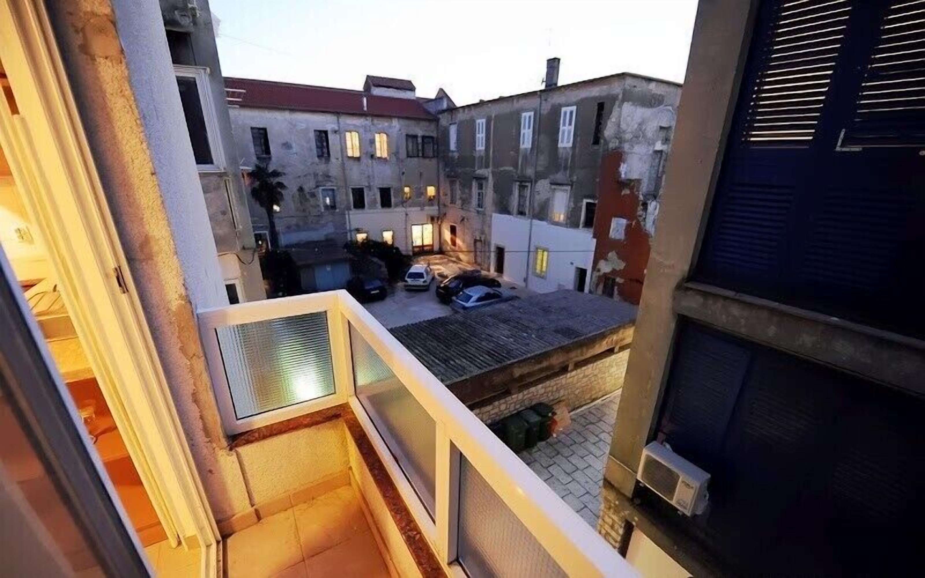 A courtyard with a view of adjacent buildings, a small parking area, and a balcony with a railing.