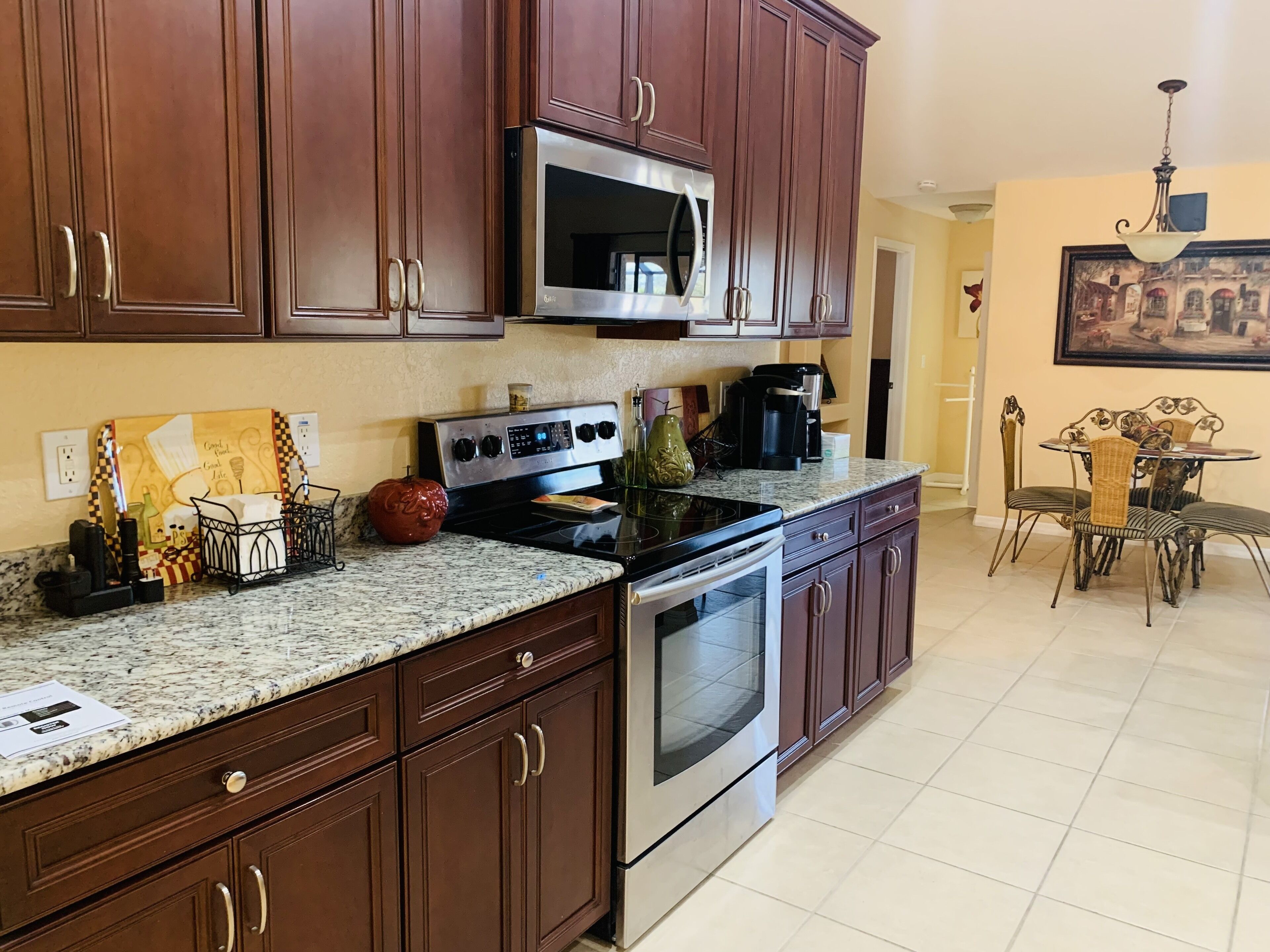 A kitchen with dark wood cabinets, a stainless steel microwave, a stove, and a coffee maker. There is a dining table with chairs in the adjacent dining area.