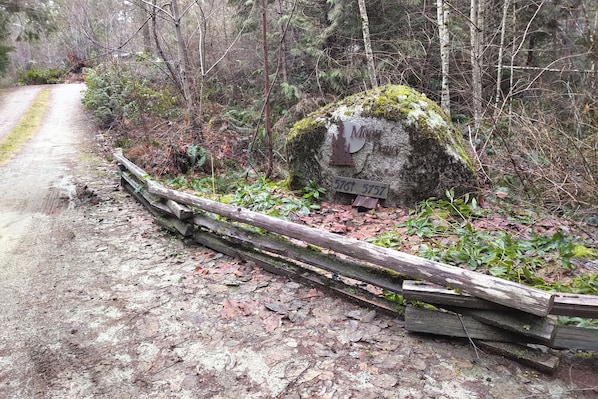A rustic wooden fence, a moss-covered rock with a sign, and a dirt path in a forested area. - The Perch at Moon Dance (Madeira Park)