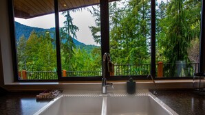 A kitchen with a large window, a sink, and a view of a forested mountain. - The Perch at Moon Dance (Madeira Park)