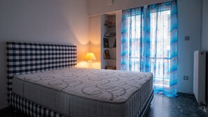 A bedroom with a checkered headboard, a bedside lamp, a bed with a patterned bedspread, a small shelf with items, and a window with blue curtains. - Modern design - Lux 1 B Apart. -15 m. from Acropolis! (Athina)