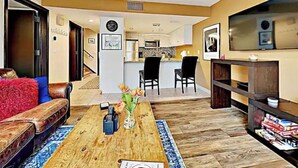 Una sala de estar con un sofá, una mesa de centro y un televisor. Se ve una cocina al fondo.