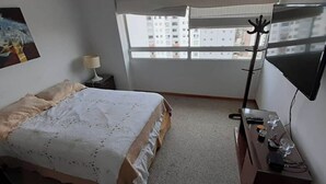 Ein Hotelzimmer mit Bett, Schreibtisch, Lampe, Fernseher und einem Fenster mit Blick auf Gebäude.
