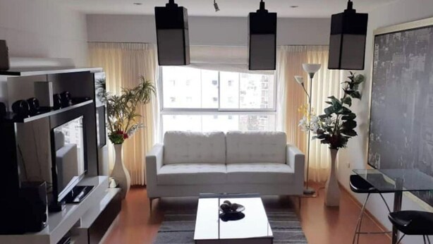 Ein modernes Wohnzimmer mit Sofa, Couchtisch und Fernseher. Zwei Pendelleuchten, ein Bücherregal und ein großes Fenster mit Vorhängen.